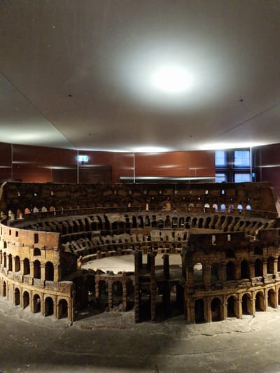 The image depicts a large, intricately detailed architectural model of the Roman Colosseum, located within an indoor exhibition space in Aschaffenburg, Germany, specifically at or in association with the Pompejanum.
The model occupies the central and foreground portions of the frame, presented from an elevated perspective looking down into its interior. It is constructed from a reddish-brown material, simulating aged brick or stone, and accurately replicates the multi-tiered arcaded structure, including visible internal hypogeum elements within the arena. The model exhibits intentional distress and partial ruin, reflecting the Colosseum's current state. It rests on a flat, light grey display platform that extends into the foreground.
The surrounding environment features a high, light grey ceiling with a prominent, bright circular light source reflecting off its smooth surface, positioned towards the upper right. The walls are a dark reddish-brown, possibly curved, forming the background. On the left wall section, a green illuminated exit sign with a white running figure pictogram is visible. In the distant background, centered horizontally, are large, multi-paneled windows displaying a cool blue light, suggesting an exterior view or distinct lighting. The overall illumination is subdued, with areas of deep shadow beneath the model's arches and within its interior, contrasting with the brighter ceiling and windows. No individuals are present in the scene.