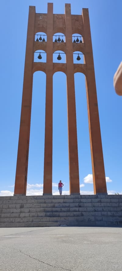 A wide-angle, low-angle shot captures the monument of the Armenian Apostolic Church in the city of Araks, Armenia. The monument, made of reddish-brown stone, is a tall, narrow tower with three arched openings. Each opening contains a row of bells. A person wearing a pink shirt and blue jeans stands on a wide, stone staircase leading up to the monument. The sky above is a clear blue with a few scattered white clouds. The foreground shows a cracked asphalt surface. The image is composed with the monument dominating the upper portion of the frame, and the staircase and person occupying the lower half.