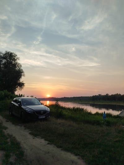 This is a horizontal, eye-level, medium shot of a dark-colored Mazda CX-5 parked on a dirt path next to a grassy riverbank at sunset. The car is positioned in the lower left quadrant of the frame, facing slightly to the right. Its headlights are on, and the license plate reads "WZY 37745". To the right of the car, tall green grass and reeds line the edge of a calm river, which reflects the warm hues of the setting sun. The sun, a bright orange orb, is low on the horizon, casting a glow across the water and the sky. The sky above is a soft blend of blue, pink, and orange, with wispy clouds. A line of trees forms a dark silhouette along the opposite bank of the river. In the foreground, the dirt path is visible, leading away from the car into the lower left corner. A partially visible blue umbrella on a stand is planted in the grass near the riverbank on the right side of the frame. The overall scene depicts a tranquil evening at a riverside location, potentially in or near the city of Topolina, Poland, given the context.