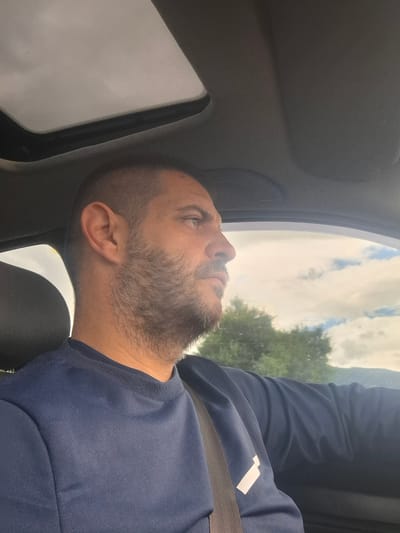 A man is captured in a profile view from the chest up, looking out of a car window. He has short dark hair and a beard, and is wearing a dark blue sweatshirt with a white geometric design on the left sleeve. A seatbelt is visible across his chest. The interior of the car, including the sunroof and the dashboard, is dimly lit. Outside, through the window, a cloudy sky and green trees are visible. The image appears to be taken in Gabarevo, Bulgaria.