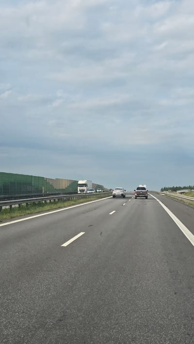 A highway scene is depicted from the perspective of a moving vehicle. The road is paved with asphalt and marked with white lines. In the distance, several vehicles are visible, including cars and a truck, traveling in the same direction. A sound barrier, made of green panels, lines the side of the highway. The sky is overcast with a pale blue hue, suggesting it is daytime with cloudy weather. The landscape beyond the sound barrier appears to be rural, with some trees and greenery visible. There are no visible people, and no text is present in the image. The overall impression is one of typical highway travel in a rural setting.