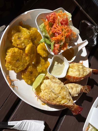 This image depicts a prepared meal presented on a large white oval plate, set on a dark brown wooden slatted table, likely in an outdoor dining environment at night in the city of Roatán, Honduras. The plate holds several components: numerous golden-yellow fried plantains (tostones) occupying the left side; three cooked lobster tails, split lengthwise with white meat and a lightly browned, possibly cheesy, topping, positioned on the right; a bright green lime wedge placed among the food items. Accompanying the main dishes are two small white, shell-shaped condiment bowls: one containing a creamy, pale yellow sauce, and another holding a translucent amber liquid. A separate small white, boat-shaped bowl, also resting on the main plate, contains a mixed green salad composed of lettuce, diced red tomatoes, and shredded orange carrots. In the lower-left foreground, a white paper napkin is wrapped around a dark object, possibly cutlery. A blurred glass object is partially visible in the upper-right background. No people are visible.