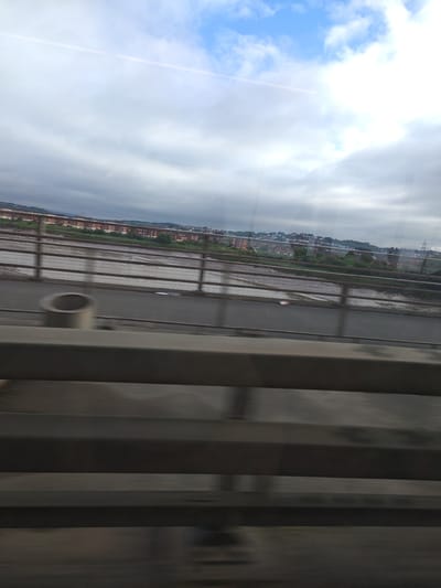 The image captures a view from a moving vehicle, likely a train or car, looking out over a wide, muddy river or estuary. In the distance, across the river, a dense urban area with numerous residential buildings, primarily brick structures with red-tiled roofs, is visible stretching along a hillside. The sky above is overcast with patches of blue peeking through the clouds. In the foreground, the blurred image of the vehicle's interior structure, including what appears to be a railing or barrier, is prominent. The location is the city of Newport, United Kingdom, with the river likely being the River Usk. The scene depicts a typical urban landscape adjacent to a river, under a partly cloudy sky, viewed from a moving perspective.