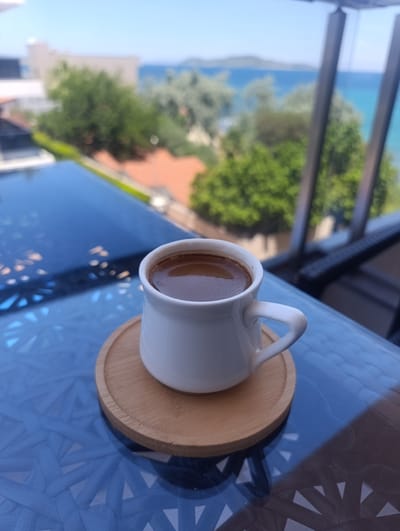 A white cup of coffee sits on a wooden coaster on a reflective blue table. In the background, out of focus, is a view of the Aegean Sea, green trees, and buildings, suggesting the location is Urla, Turkey. The sky is clear and blue. There are no people visible in the image.