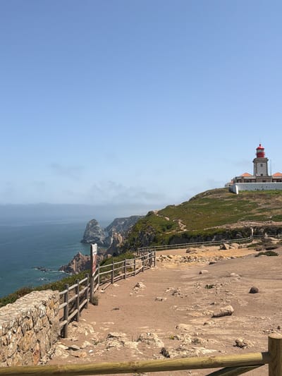 A lighthouse stands on a grassy hill overlooking the ocean near Colares, Portugal. The lighthouse is white with a red roof and sits on a white building. A wooden fence runs along the edge of the cliff, separating the grassy hill from a sandy path. The ocean is a deep blue, with some rocks visible near the shore. The sky is clear and blue, with a few clouds in the distance. The area around the lighthouse appears to be a tourist attraction, with a signpost visible near the fence. The overall scene is serene and picturesque, with the lighthouse serving as a prominent landmark against the natural beauty of the coastline.