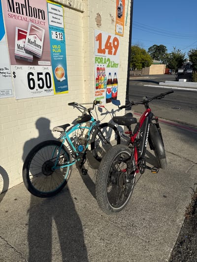 The image displays an outdoor scene in what appears to be an urban or suburban setting, possibly in Rio Linda, United States. Two bicycles are leaning against a light-colored brick building on a sunny day, indicated by the bright blue sky and distinct, elongated shadows suggesting either early morning or late afternoon.
One bicycle is a red and black electric bike, with "MOKWHEEL OBSIDIAN" visible on its frame, featuring robust, knobby tires. Leaning in front of it is a light blue bicycle that has been customized with a small engine kit, including a motor mounted near the pedals and a fuel tank on the top tube, making it a motorized bicycle. Both are parked on an asphalt surface next to the building.
The building's wall is covered with several advertising posters. One poster advertises RC Cola and other sodas for "$1.49 FOR A 2 Liter," mentioning "AMAZING FLAVOR" and "Cherry." Below this, a Marlboro Special Select cigarette advertisement promotes a "SAVE 650 WHEN YOU BUY 2" deal and includes a health warning. Another sign advertises "VELO PLUS" nicotine pouches, offering to "SAVE $5.12 WHEN YOU BUY 2" and carrying a warning about nicotine being an addictive chemical. In the background, trees, power lines, and distant structures are visible under the clear sky. No people are present in the image.