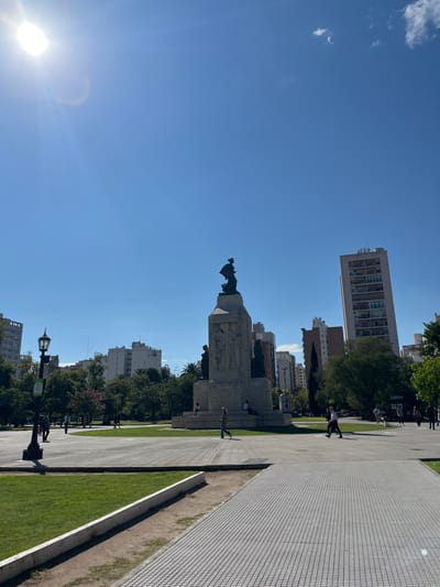 The image depicts a sunny day at Plaza Rivadavia in Bahía Blanca, Argentina. A large monument with a statue on top stands prominently in the center. Several people are strolling through the plaza, some walking along the paved walkways and others near the grassy areas. The plaza is surrounded by buildings, including a tall apartment complex. The paving is a white block grid. The grass looks well-manicured. A lamppost is visible in the foreground. The sky is blue with only a few clouds.