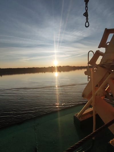 The image captures a serene sunset over a body of water, likely a lake or a wide river, with a treeline forming a dark silhouette in the distance. The sun, a bright orb, is low on the horizon, casting a golden glow across the sky and reflecting on the water's surface, creating shimmering ripples. Two distinct contrails are visible in the sky, suggesting recent air traffic. In the foreground, the edge of a boat or ferry is visible, with a dark green deck and some metallic structures painted in a light yellow hue. A dark chain hangs from the upper right portion of the frame. The scene is set during the golden hour, emphasizing the calm and tranquil atmosphere of the location in Bol'shaya Stepanovskaya, Russia.