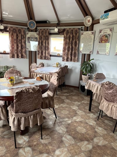 The image depicts the interior of a dining area, likely a cafe or restaurant, in Русский Турек, Russia. The space is characterized by a warm, traditional aesthetic. Several tables are set with tablecloths and chairs covered in decorative fabric. One table in the foreground is round and dark wood, set with placemats, salt and pepper shakers, and a packet of mustard. Another table in the midground is rectangular and covered with a white tablecloth.
The walls are adorned with framed posters, one featuring an image of food with Cyrillic text and another with a recipe for "Pelmeni USSR." Drapes with a patterned design hang over windows, and decorative plates are mounted on the ceiling beams. A potted plant sits on a stand near a wall-mounted air conditioner. The floor is tiled with a geometric pattern in shades of brown and beige. The overall impression is one of a cozy and culturally themed dining establishment.