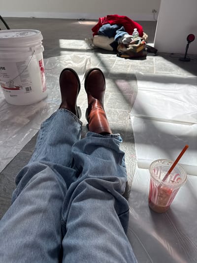 The image shows a person sitting on a plastic drop cloth wearing blue jeans and brown boots. A white bucket is to the left, and a clear plastic cup with a straw is to the right. In the background, a pile of clothes and a desk lamp are visible. The location appears to be indoors, possibly a room being prepared for painting or renovation. The city of Town of Red Hook, United States of America is only a best guess. No other specific details are available to pinpoint an exact location within the city.