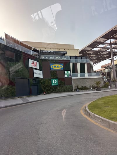 A side view of a modern shopping center is captured in this image, located in Urbanización La Cenia II, Spain. The building features a prominent green wall adorned with various store signs, including H&M, Cortefiel, IKEA, and Deichmann. Further up, a sign reads "LIZARRAN PINCHOS, TAPAS Y MUCHO MÁS." A portion of the building has a distinctive cantilevered roof structure. In front of the building, a paved walkway runs alongside a grassy area. Several individuals are seated on a low wall in front of the building, while others are standing near the entrance to another section of the shopping center, which has a sign that reads "Tapas & Beer." The scene is framed by a road with a yellow line marking the edge, curving towards the right side of the image.