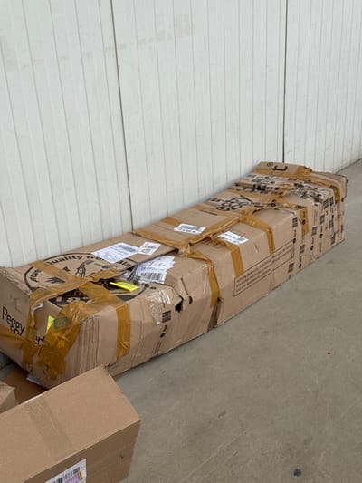 In Bad Säckingen, Germany, two large cardboard boxes are positioned outdoors on a concrete surface. The primary box, which is long and narrow, lies horizontally against a white, ribbed wall. It is heavily taped with brown packing tape, some of which is torn. Shipping labels and barcodes are visible on its surface, along with printed text that includes "Quality from Bla" and "Peggy". There are also several instances of "dpd" branding visible on the box. A second, smaller cardboard box is situated in the lower left foreground, partially obscuring the main box. It also appears to be a shipping box, with a white label bearing a barcode. The lighting suggests it is daytime.