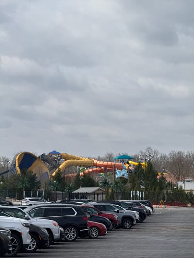 The image shows a parking lot filled with cars, with a water park visible in the background. The water park, identifiable as Zoombezi Bay based on the distinctive water slides, is located in Powell, Ohio, United States of America. The weather appears overcast. There are no people visible in the image. The prominent features are the water slides of the park and the cars in the parking lot. There are no interactions or actions taking place in the photo.