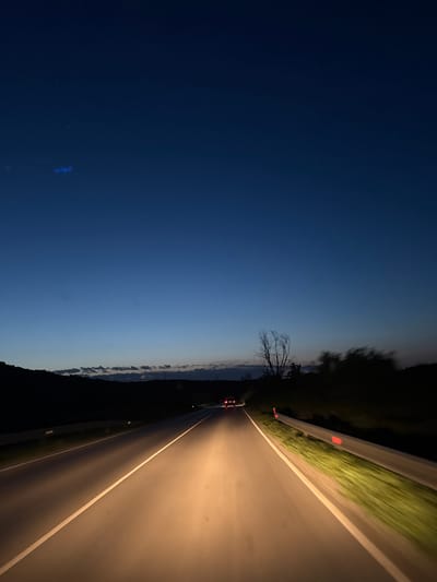 The image presents a night scene on a road in Zdravets, Bulgaria. The sky transitions from a deep blue to a lighter blue near the horizon. A dark treeline runs along the horizon with a bare tree visible in the center. On the right, a brightly lit road stretches into the distance, illuminated by the headlights of a car ahead. A guardrail and roadside vegetation are visible along the road's edge.