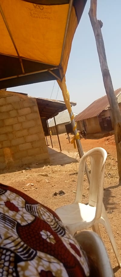 The scene is an outdoor, sunlit area in Kasuwan Magani, Nigeria, characterized by dry, reddish-brown earth. In the immediate foreground, the lower right quadrant reveals a person's lap covered by a patterned fabric featuring red, white, brown, and black motifs. To the right of this, a white plastic chair stands on the dirt. Overhead, a yellow fabric canopy, likely part of a tent structure, is visible, supported by metal poles. Faint text, including "KD" and "ORDER YOURS", is discernible on the canopy's underside.
In the midground, a structure constructed from light-colored concrete blocks with a corrugated metal roof supported by wooden posts stands to the left. A metal pole, partially wrapped in yellow fabric, is prominent, alongside a rough wooden pole. The ground is consistently dusty and dry, with minimal vegetation and scattered debris.
Further in the midground and background, several figures, appearing to be children, are observed on the dirt path between buildings. Multiple single-story residential or commercial structures with corrugated metal roofs, varying in color from silver-grey to rusty brown, line the distant perimeter. The sky above is clear and light blue, indicating bright daylight.
