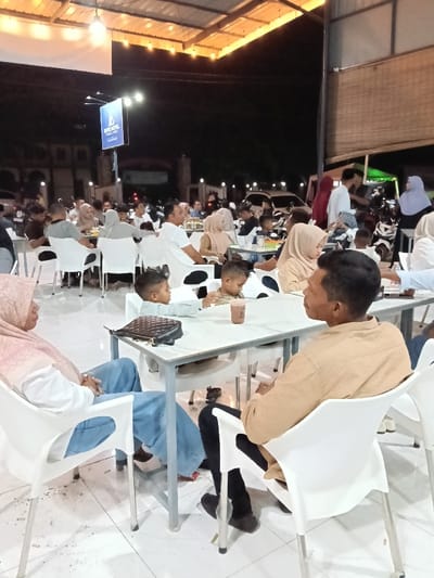The image captures a nighttime scene at a bustling outdoor or semi-covered eatery in Kampongbaro, Indonesia. Numerous individuals, including adults and several children, are seated at white rectangular tables with metal frames, utilizing white plastic chairs.

In the foreground, a woman wearing a light pink headscarf and light blue pants is seated on the left, looking generally forward. To her right, a man in a tan long-sleeved shirt and dark pants is seated, facing left. Between them, at a table, two young boys are present; one in a light blue long-sleeved shirt, the other in a patterned shirt, with a dark brown beverage cup and a black quilted handbag resting on the table surface.

The environment is illuminated by overhead string lights and exposed bulbs affixed to a corrugated metal roof structure. In the mid-ground and background, other patrons are engaged in conversation and dining. A prominent blue sign for "BUE HOTEL FAMILY STAY" is visible in the background, indicating nearby commercial establishments. Vehicles, including cars and motorbikes, are discernible in the distance, suggesting proximity to a street or parking area. The floor is covered with light-colored tiles, and a bamboo-like roller blind is partially visible on the right side. The overall scene depicts a public gathering place, well-lit despite the night setting.