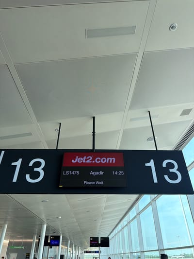 The image shows a sign at an airport in Uttlesford, United Kingdom, indicating gate 13 for a Jet2.com flight LS1475 to Agadir, departing at 14:25. The sign displays "Please Wait". The airport has a modern design with a glass ceiling allowing natural light to enter. Other gate signs can be seen in the distance, along with support columns. The wall is composed of rectangular panels with a perforated texture.