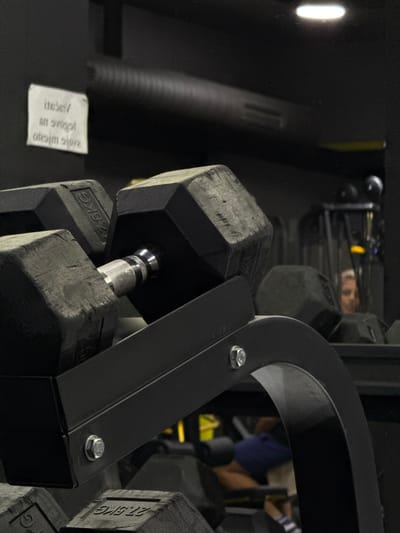 A close-up captures a rack of black, hexagonal dumbbells in a gym setting. The rack is made of black metal with visible bolts. Reflections and other gym equipment are faintly visible in the background. The dumbbells range in weight, with one labeled "7.5KG" and another "27.5KG." A partially obscured white tag with printed text is near the rack.