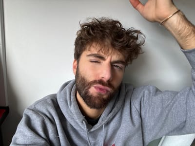 This image depicts a single male individual in a close-up, chest-up portrait, located indoors in La Garenne-Colombes, France. The subject has brown, curly hair and a full brown beard. He is wearing a grey hooded sweatshirt. His right arm is raised, with a thin, gold-colored bangle present on his right wrist; the hand is positioned above the frame. The individual's left eye is closed or tightly squinted, while the right eye is partially visible. His lips are pursed into a 'duck face' expression, directed towards the camera. The background consists primarily of a plain, light-colored wall. A small segment of a red object with a white border is visible on the far left edge of the frame, and a textured white object is partially visible on the lower right.