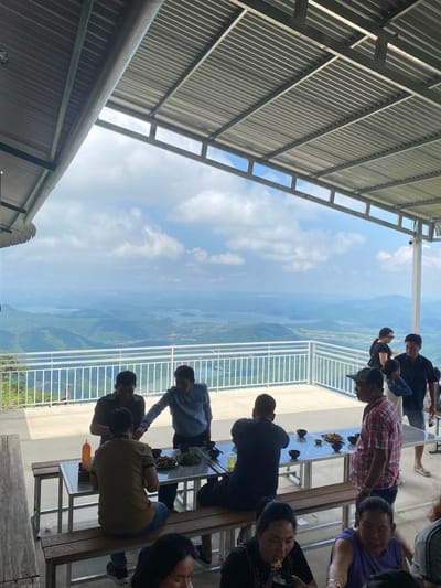 The image depicts a group of people dining at an outdoor restaurant with a panoramic view of a mountainous landscape featuring bodies of water. The setting appears to be a terrace or balcony overlooking a vast valley. The architecture suggests a roofed area, possibly a pavilion, supported by metal structures, offering shade. Several individuals are seated at tables, some engaged in eating and conversation. The food appears to be served in bowls, and drinks are present on the tables. In the background, lush green mountains and expansive lakes or rivers are visible under a cloudy sky. The overall impression is that of a scenic dining experience. The location in the city of Phan Son, Vietnam is consistent with the visual elements of a mountainous, water-rich landscape.