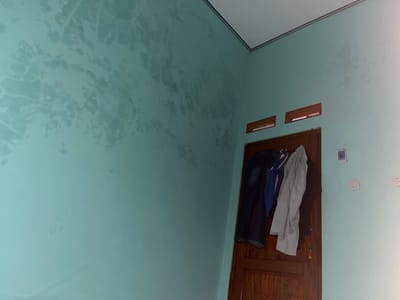 A corner of a room in Meteseh, Indonesia, shows a light teal-colored wall with water stains forming irregular, lighter patterns. A wooden door is visible, with a pair of dark blue jeans and a gray jacket hanging from it. Above the door are two rectangular wooden ventilation grates. On the wall to the right of the door, there are two white electrical outlets or light switches. The ceiling is white with a thin dark trim along the edge. The lighting in the room suggests it is daytime, with light casting shadows.