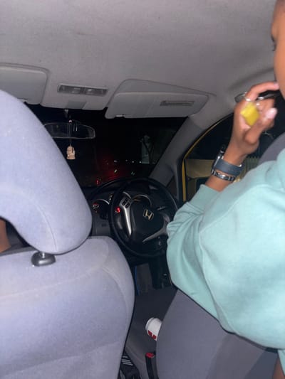 Inside a car at night, someone is cleaning the ceiling with a yellow sponge. The person wears a light blue sweatshirt, grey leggings, and a watch. The dashboard with the steering wheel, the seats, and the rearview mirror are visible. Outside the car, there are lights visible through the windows.