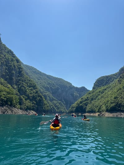 The image depicts a group of people kayaking on a wide, calm, turquoise lake, with steep, verdant mountains rising on either side. The sky is clear and blue. The kayaks are of various colors, including orange, yellow, and red, and several individuals are actively paddling. The mountainsides are densely covered in green trees, with some rocky outcrops visible. In the foreground, the water has a rippling texture, reflecting the clear sky. The scene appears to be a natural landscape, possibly a canyon or a fjord, in Bezuje, Montenegro, a region known for its stunning natural beauty and outdoor recreational activities like kayaking.