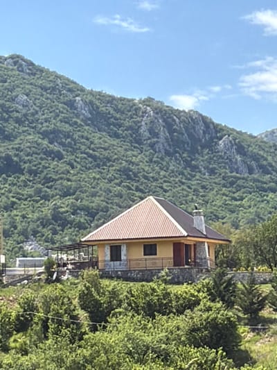 A wide shot captures a yellow house with a terracotta roof nestled on a hillside in Vrela, Montenegro. The house, featuring stone accents and a prominent chimney, is surrounded by lush greenery and overlooks a densely forested mountain range under a clear blue sky with scattered clouds. A small patio area is visible on the left side of the house, and a satellite dish is perched on the hillside. The foreground is filled with dense trees and vegetation.