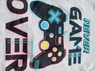 This is a close-up, vertically oriented image of a light-colored t-shirt with a graphic design. The design features a black and brightly colored illustration of a video game controller. The controller is stylized with neon pink and cyan outlines and accents, including a cyan "X" button, circular buttons with yellow centers and pink outlines, and various other geometric shapes. To the left of the controller, the word "OVER" is printed vertically in large, bold, black letters with neon pink outlines. To the right of the controller, the words "GAME NEVER" are printed vertically, also in large, bold black letters with cyan outlines. The t-shirt fabric has a visible texture with some slight wear and discoloration. The overall aesthetic of the graphic suggests a gaming theme, possibly related to retro or arcade games, with a vibrant and energetic style. There are no people, specific scenery, or weather conditions visible. The text present is "OVER" and "GAME NEVER". The location context provided is Oleksandriia, Ukraine, but this cannot be visually determined from the image itself.