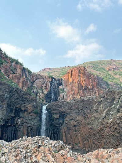 The image shows a waterfall cascading down a rocky cliff face. The rocks are layered and appear to be composed of dark volcanic rock with some lighter, reddish-brown rock formations interspersed. Vegetation, consisting of small shrubs and grasses, grows in crevices on the cliff face. A thin wire is visible stretched horizontally across the rocks in the middle ground. The sky above is blue with scattered white clouds. The overall scene suggests a natural landscape, likely a remote or mountainous area. The lighting indicates it is daytime, possibly late morning or early afternoon, with bright sunlight illuminating parts of the cliff face and creating shadows in others. There are no visible people or text in the image. The orientation of the image is portrait, with the cliff face running diagonally from the bottom right to the top left. The presence of a waterfall and the rugged, natural terrain are the primary subjects.