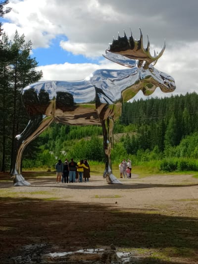 This is a full-shot, eye-level, outdoor, vertical photograph of a large, polished metallic moose sculpture in Atna, Norway. The sculpture is highly reflective, mirroring the sky, clouds, trees, and some people in the background. The moose is positioned on a dirt path, with its front left leg extended forward and its hind legs bent. Its large antlers are prominent.
In the background, a group of approximately 6-8 people are standing on the path, looking towards the sculpture. Some appear to be adults, and at least one appears to be a child. The environment consists of a wooded area with numerous evergreen trees forming a dense backdrop behind the sculpture and the people. The sky is partly cloudy, with patches of blue sky visible between the clouds. The foreground of the image is a dirt path with some muddy patches and scattered stones, partially in shadow.