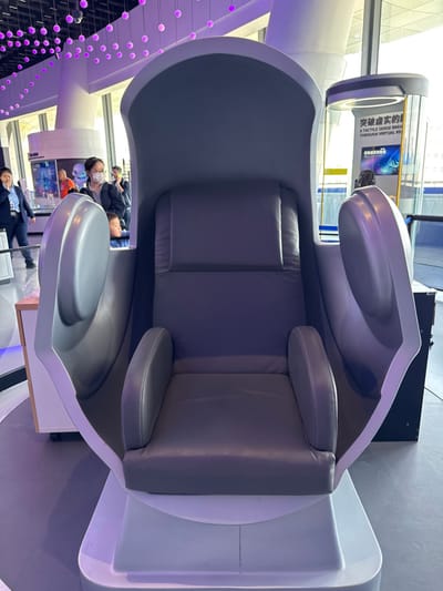 An unoccupied, futuristic-style recliner chair, predominantly white with gray upholstered seating, occupies the foreground of an indoor exhibition hall in Guangming District, China. The chair features a high back, integrated armrests, and partial side enclosures, all supported by a white, low-profile base. The floor is a smooth, dark gray. Overhead, numerous small, spherical lights emit a purple glow from the ceiling. In the background, multiple individuals are present: to the far left, a person in dark professional attire appears to be speaking on a mobile phone. A woman wearing a white face mask stands with a young child near a digital display exhibiting blue graphics. To the right, a tall, cylindrical display unit features text in Chinese and English, stating "A TACTILE SENSE BREATH THROUGH VIRTUAL REALITY." Large windows providing natural light are visible beyond the displays. No direct interaction with the main chair is occurring.