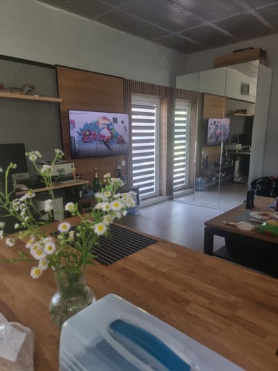 The image depicts an indoor scene, likely a living room or dining area in a home in Szczaki, Poland. The room is furnished with a large wooden table in the foreground, adorned with a bouquet of small white and yellow flowers in a glass vase. The table also has a white plastic object with a blue section on its right. In the background, a wall features large mirrored closet doors, reflecting the room and showing more of the interior. A television screen displays a colorful animated image. There are blinds on the windows, suggesting it's daytime. The overall ambiance appears relaxed and domestic.