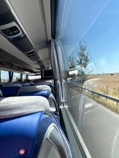The image shows the interior of a bus, with a view through the large windows. The perspective is from a seat looking down the aisle and out the side and overhead windows. The bus appears to be in motion, as indicated by the blur of the scenery outside.
The subjects are the seats within the bus, which are upholstered in white and blue. The aisle runs between rows of these seats. Outside the window, a dry, grassy landscape is visible under a clear blue sky. There are some trees and a fence line along a road.
The activity taking place is travel. The time of day is clearly daytime, with bright sunlight streaming through the windows, suggesting it's likely midday or afternoon. The weather appears to be fair and sunny.
Notable details include the ribbed texture on the interior panels above the seats, and the air vents with circular blue logos. The overhead windows are covered with a protective plastic film. The seats have a modern design.
There is no visible text in the image. The location context is Shishmantsi, Bulgaria, which aligns with the dry, rural landscape and sunny weather. No specific emotions are conveyed, as the image focuses on the interior and exterior environment of the bus.