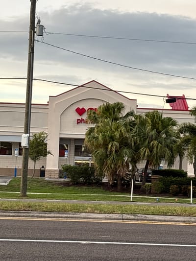 A CVS Pharmacy is visible from the exterior, with its red heart logo and "pharmacy" signage displayed prominently on the building's facade. The building features a light-colored exterior with a red trim along the roofline. In front of the pharmacy, palm trees and landscaping are present. A utility pole with a transformer is situated to the left of the building, and power lines cross overhead. The scene appears to be in a commercial area, with a road and sidewalk in the foreground.