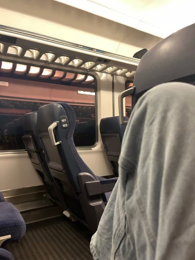 The image displays the interior of a passenger train carriage. The location in the city of Klikach, Bulgaria, cannot be confirmed by any visual information within the frame.
In the foreground, the lower torso and legs of an individual wearing light blue denim or chambray-like fabric are partially visible, indicating a seated position directly in front of the camera's perspective. Ahead, two rows of dark blue fabric train seats with grey plastic backs are visible, oriented away from the camera. The seat closest to the center displays "101" stacked above "103" in white text on its headrest, and has a silver-colored grab handle attached to its upper back.
Along the side of the carriage, multiple rectangular windows show a blurred, warm reddish-brown exterior, suggestive of a station platform at night or dusk, with reflections of the train's interior also present. Above the windows, light-colored overhead luggage racks with an open-lattice design extend along the ceiling. The floor is dark and features a ribbed texture. Artificial lighting within the carriage casts a dim, uniform illumination. No explicit actions or interactions are discernible.