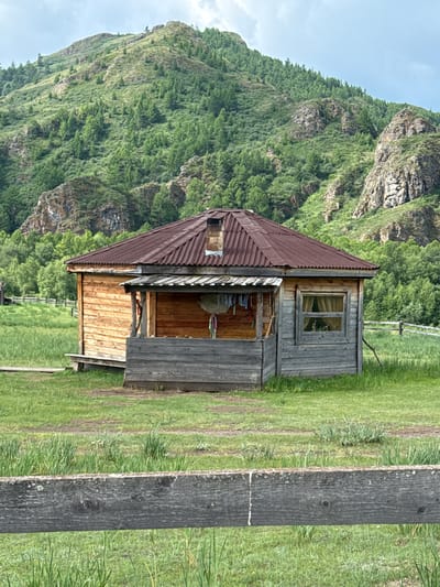 The image depicts a rustic wooden cabin situated in a grassy field with a lush, green mountainous backdrop. The cabin has a corrugated metal roof and a porch area. Several items are hanging on the porch, possibly laundry. A window with curtains is visible on the right side of the cabin. The foreground shows a weathered wooden fence. The environment suggests a rural or remote setting. The location is identified as Kazanovka, Russia.