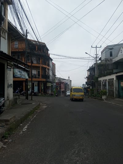 This image depicts a street scene in the city of Houaythat, Laos, under an overcast sky. The asphalt road, dark in tone, occupies the central and lower portions of the frame. A concrete curb and uneven sidewalk with sparse vegetation run along the left side.
On the left, a light-colored building in the foreground shows a dark open doorway and graffiti, partially obscured by a faded banner. Further along the left, a multi-story building with an orange-brown facade features multiple balconies with black railings, and storefronts on the ground level, some displaying "SALON" signs. A dense network of black utility cables is strung overhead, connecting to poles and crossing the street, particularly prominent above this building. Several motorcycles are parked along the left sidewalk; one individual is visible near a blue motorcycle.
In the center of the road, a yellow mini-van, possibly a utility vehicle, travels towards the viewer. White passenger vehicles are visible further down the street behind it.
On the right side of the road, various buildings line the street. The foreground includes low-rise structures, some with metal gates and potted plants, and signs indicating businesses such as "Chappy" and "DION SALON." A grey-sided building with a single rectangular window is also present. A utility pole with a street lamp extends over the road, and another utility pole with a transformer is visible in the mid-ground.
In the distant background, a white structure resembling a minaret is visible, positioned centrally. No overt actions or interactions between individuals are discernible beyond the yellow van's movement and the single person near the motorcycle. The overall environment appears urban and moderately weathered.