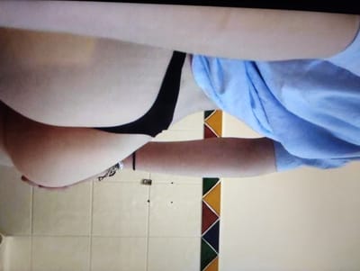 The image is a close-up, horizontal shot of a person lying down, with their body angled from the bottom left to the top right. The person is wearing a light blue, long-sleeved garment on their upper body and black underwear that is visible around the waist and buttocks. A bracelet with charms is visible on the person's left wrist.
The background consists of a tiled wall with a decorative strip featuring triangular shapes in yellow, red, dark blue, and green. The lighting appears to be artificial and comes from the front.
The scene is intimate and focuses on a portion of the person's body. The overall mood is subdued, with no strong indicators of specific emotions or a particular event. The location context provided (Brashlen, Bulgaria) is not directly discernible from the visual elements of the image itself. There is no visible text in the image.