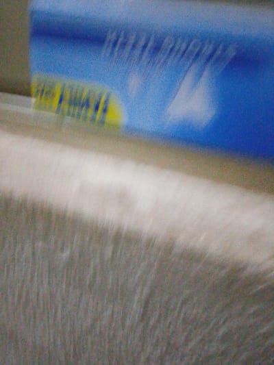 The image displays an extremely blurred and out-of-focus scene, presenting as an abstract composition. The upper portion, roughly one-third of the frame, is dominated by a vibrant blue field containing highly distorted yellow and blue markings. These markings are indistinct due to severe blur, but the yellow element loosely resembles the letters "UBA," potentially indicating a reference to United Bank for Africa. Within the blue field, faint, wavy lighter blue or white streaks are also present. The lower two-thirds of the image consist of a granular, textured field ranging from off-white to a muted greyish-brown, representing an indistinct surface. No individuals, specific objects beyond the abstract markings, or actions are discernible. The overall environment is obscured by the extreme blur, but based on the "UBA" hint, the scene may depict a commercial or banking environment located within the city of Abatete, Nigeria.