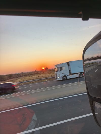 The image captures a sunset scene on a highway, possibly near Torrent, Spain, as indicated by the distinct silhouette of buildings and structures in the distance and the golden hues of the sky. A white semi-trailer truck is in motion on the highway, its trailer a large, reflective surface. To the left, a dark-colored car is also traveling, its taillights blurred by motion. The foreground of the image is dominated by the reflection of a person wearing a red shirt and grey clothing, visible on what appears to be a window or a reflective surface. The highway itself has multiple lanes marked with white lines. In the distance, a bright orange sun is setting, casting a warm glow across the landscape, which features dry vegetation and scattered trees. The sky transitions from a bright orange near the horizon to a lighter blue at the top of the frame, with faint contrails visible. A bridge or overpass can be seen in the reflection of the side mirror on the right, with other vehicles on the road below it.