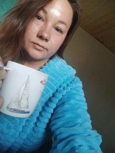 A woman is holding a white mug with a sailboat design in her left hand. She is wearing a plush blue robe with a textured pattern. The woman has long, brown hair and is looking towards the camera with a neutral expression. Her face is lightly powdered and she has freckles. The background is out of focus, but appears to be an indoor setting with a sloped wooden ceiling and a plain wall. There is no indication of the location being in the city of Вазерки, Russia.