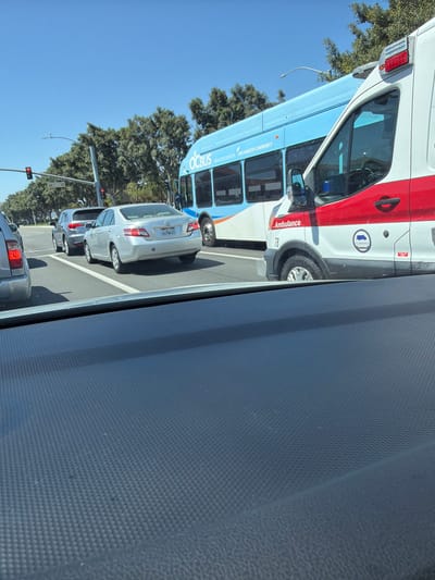The image shows a traffic scene in Irvine, California, USA, on a clear day. The view is from inside a vehicle, looking forward through the windshield. In the foreground, the textured dashboard of the vehicle is visible. In the middle ground, there are several vehicles in traffic. To the right, a white ambulance with red and white detailing is partially visible. Next to the ambulance, a blue OC Bus is seen. In front of the bus and ambulance, there are several passenger cars, including a silver sedan with license plate 6LPW620, and a gray SUV. Traffic lights and street signs, including one that reads "Hubble," are visible above the vehicles. The background features trees and a clear blue sky. The vehicles appear to be stopped or moving slowly in traffic.