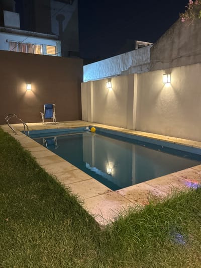 A swimming pool, bordered by grass, is situated beside a building. The pool reflects light fixtures on the building's wall, and a yellow ball floats in the water. A blue chair sits near the pool ladder. The building has exterior lights and windows.