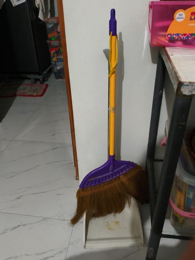 The image depicts a broom and dustpan placed on a tiled floor indoors. The broom has a yellow handle with a purple plastic attachment at the top, and a yellow string loop is tied around the handle. The bristles of the broom are brown. A white dustpan is positioned beneath the broom. In the background, a portion of a black refrigerator is visible on the left. To the right of the broom, a metal shelf unit with black legs is present, holding a pink plastic container with colorful markings and text that includes "50 pieces." The floor is white with grey veining. The overall scene appears to be a residential or storage area. The presence of household cleaning tools suggests a domestic environment. The image was taken in Cavite City, Philippines, but the specific location within the city cannot be determined from the visual information provided.