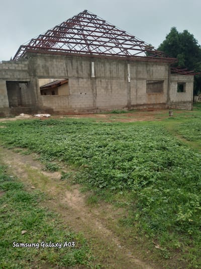 The image depicts an unfinished building with a red metal roof structure in Doka-Tofa, Nigeria. The building is constructed from gray concrete blocks, and its roof is exposed, revealing the metal framework. Openings for windows are visible, some with boarded-up sections. A second, smaller structure with a brown roof is partially visible inside one of the openings. Surrounding the building is an overgrown area with green vegetation and a dirt path. Two white sacks are placed on the ground near the building. In the background, a large green tree stands tall. The sky is overcast and gray. The lower left corner of the image displays the text "Samsung Galaxy A03".