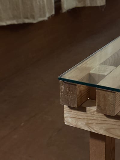 The scene, located in Suginami, Japan, displays a close-up, angled view of the corner of a table. The table features a clear glass top with visible edges, resting on a base constructed from light-toned, rough-hewn wooden blocks and planks. The wood exhibits distinct grain patterns and some surface texture irregularities, suggesting a raw or industrial aesthetic. A single vertical wooden support post is partially visible beneath the table's structure on the lower right. The background is out of focus, revealing a dark brown floor surface that occupies the left half of the frame. In the upper left background, a blurred, light-colored fabric object, possibly a curtain or upholstery, is visible. No individuals, discernible actions, or interactions are present in the image.