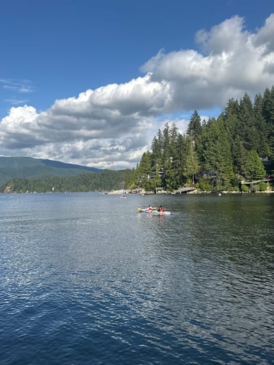 This image depicts a serene lake scene in the District of North Vancouver, Canada, under a partly cloudy sky. In the foreground, the deep blue water is textured with gentle ripples, reflecting the light. A two-person kayak is positioned in the middle ground, with two individuals, one in a red top and the other in a white top, paddling. Further back, a forested hillside rises, densely packed with tall evergreen trees. Several houses are nestled among the trees along the shore, some featuring elevated walkways and docks. A long pier extends into the water from the right side of the frame. In the distance, a range of mist-shrouded mountains forms the backdrop. The overall impression is one of natural beauty and peaceful recreation.