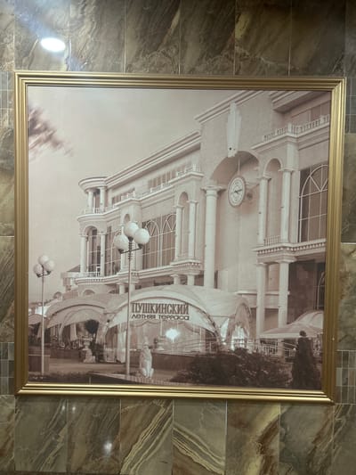 The sepia-toned photograph, framed in gold, depicts a grand building with classical architectural elements, including columns, arched windows, and balconies. A large clock is visible on the facade. In the foreground, there are illuminated street lamps and awnings over an outdoor seating area, suggesting a restaurant or cafe. The text visible on one of the awnings reads "ПУШКИНСКИЙ ЛЕТНЯЯ ТЕРРАСА", which translates to "Pushkinsky Summer Terrace". This indicates the location is the Pushkinsky Summer Terrace in Kursk, Russia. Several figures are visible, including what appear to be statues of lions and people walking. The building is set against a cloudy sky. The photograph is mounted on a wall covered with marbled tiles.