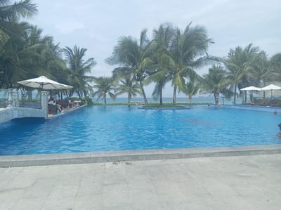 This image depicts a swimming pool area with palm trees and the ocean in the background, suggesting a tropical resort location. The pool is filled with clear blue water. Several people are visible in and around the pool. Some are swimming, while others are relaxing on chairs under umbrellas. The architecture suggests a modern resort. The overall scene is tranquil and suggests a vacation setting.